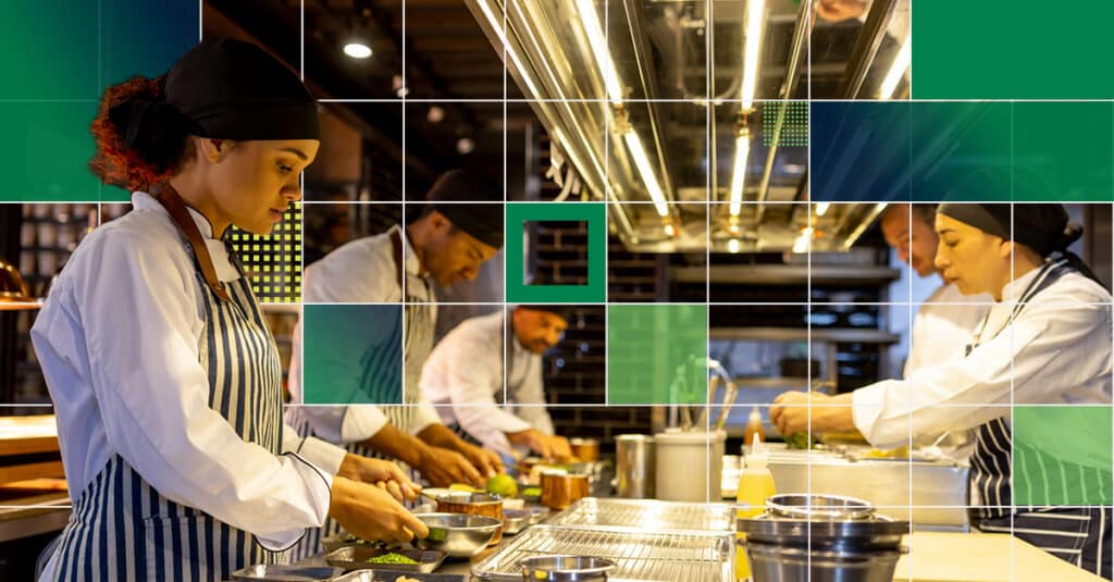 Chefs in white uniforms and striped aprons work at a busy studio kitchen, preparing plated dishes.