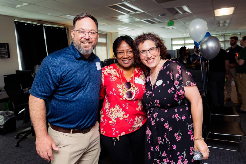 Three coworkers posing and smiling for a photo at an office celebration, with name badges and balloons in the background.