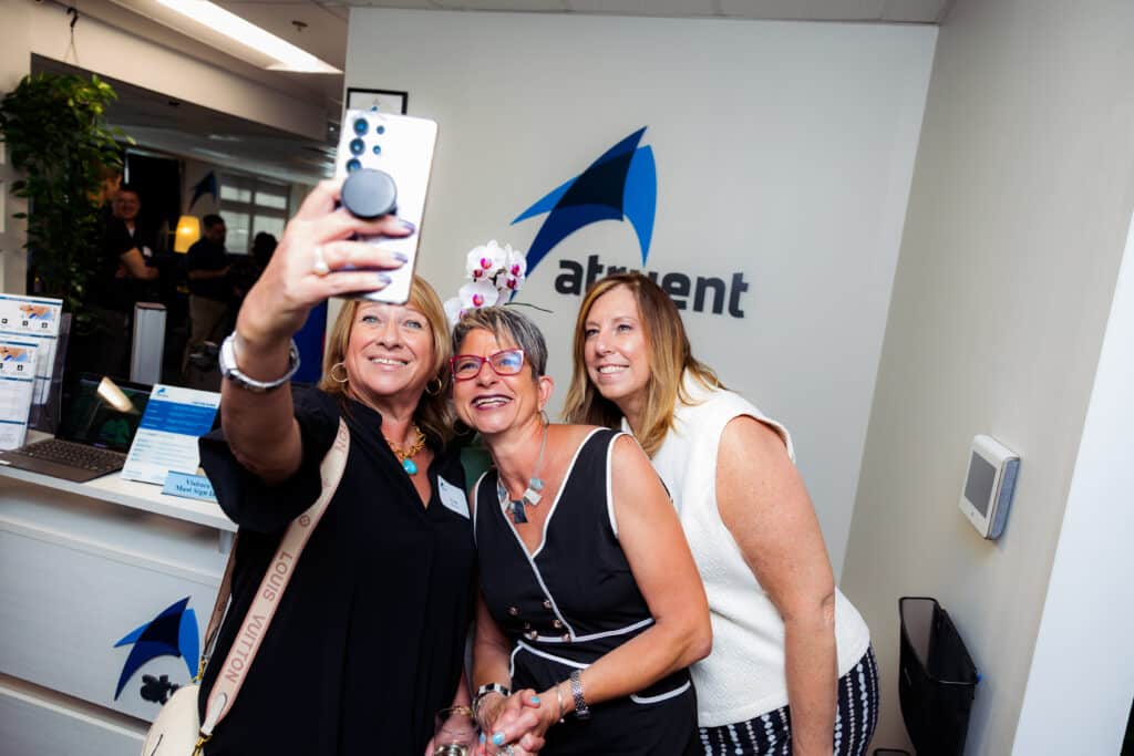 Three women smile for a selfie at a reception desk in an office, one holding a white smartphone toward the camera.