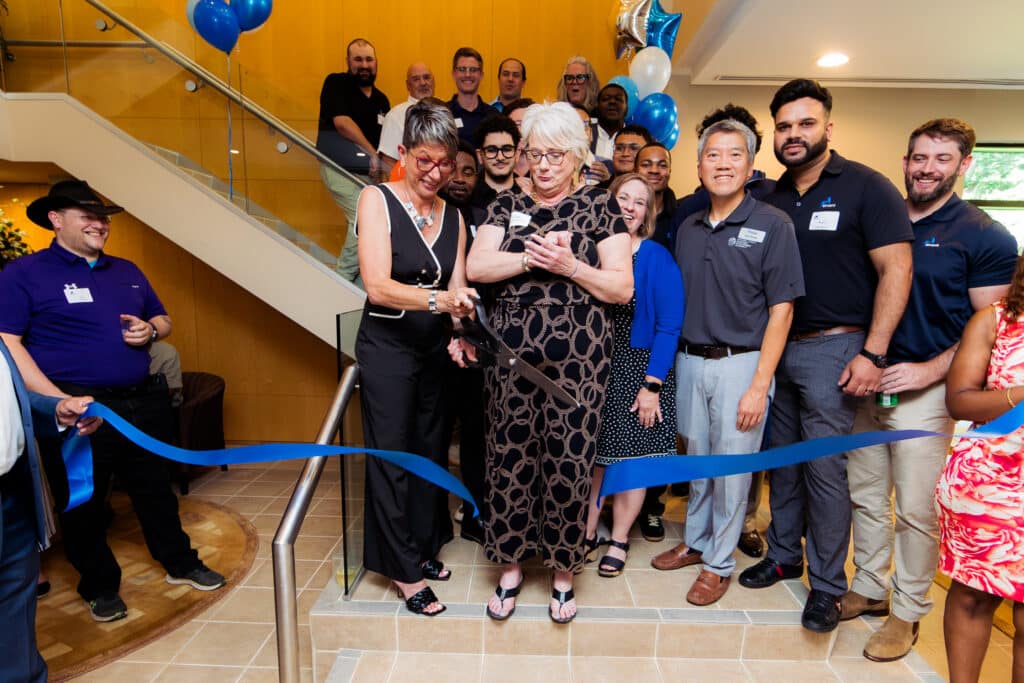 Group of people at a ribbon-cutting ceremony indoors, two women in front cutting a blue ribbon as others smile nearby, balloons visible in background