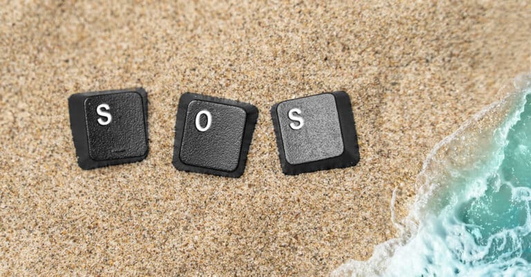 Three black keyboard keys spelling SOS lie on sandy beach with the turquoise sea nearby.