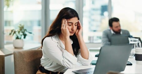 Woman at her desk rubbing her temples in a stressed moment at a laptop in an office; another coworker sits blurred in the background.